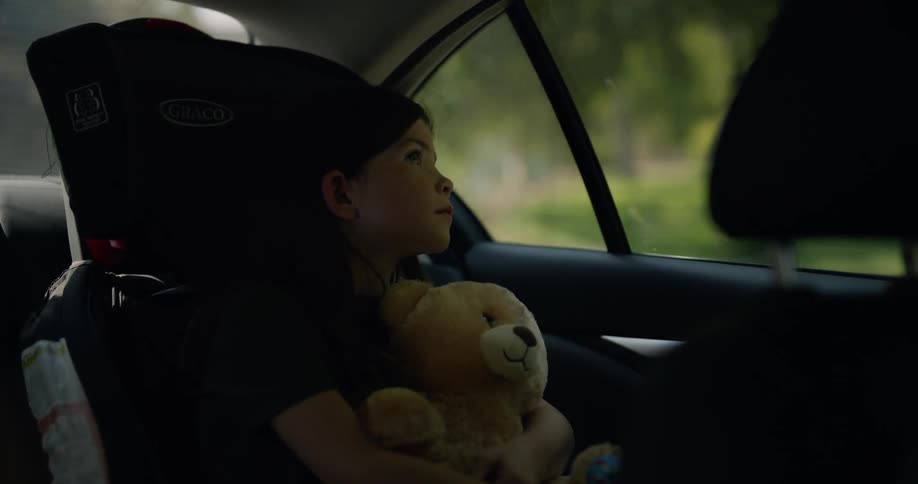 Girl with black shirt holds teddy bear and sits in safety car chair