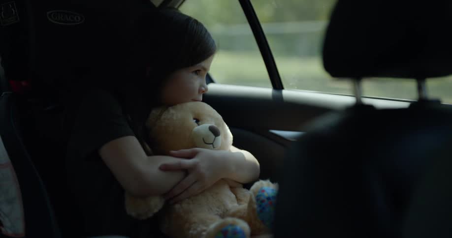 A man driving a car smiles as he looks into the mirror to see the young girl hugging a teddy bear in the back seat