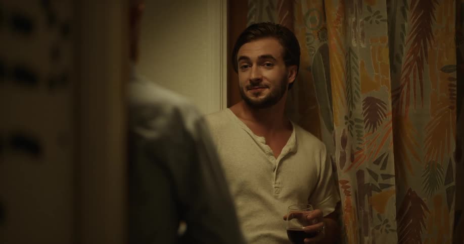 Young man reclined on a wall holding a glass of wine talks to a friend at home