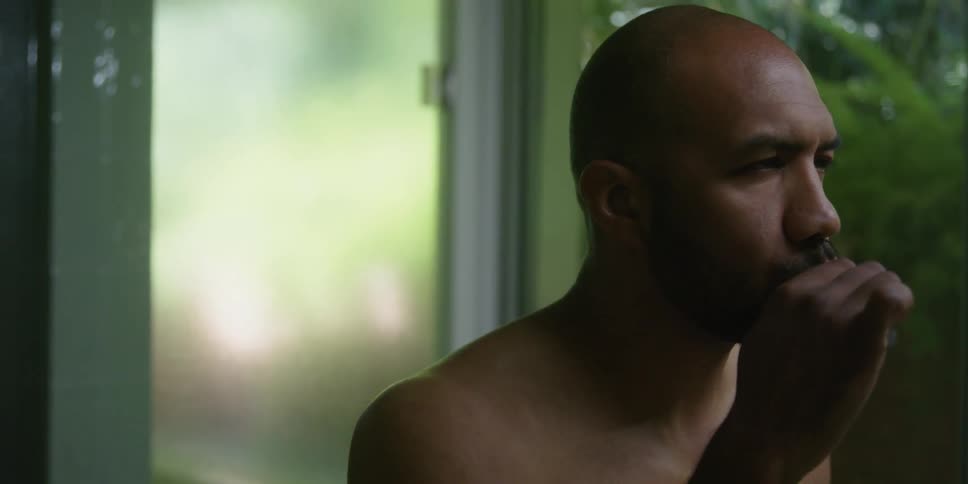 Shirtless bald man brushes his teeth in front of a window at morning