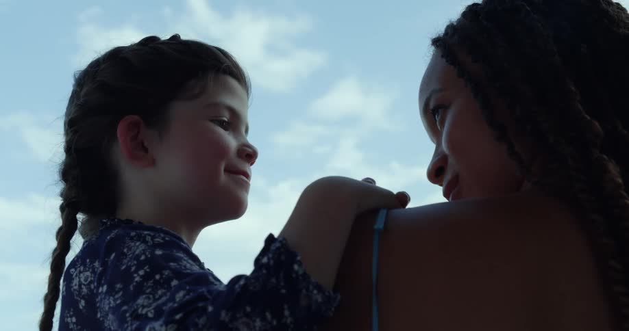 Woman with dreadlocks makes eye contact with girl with braids and blue dress smiling