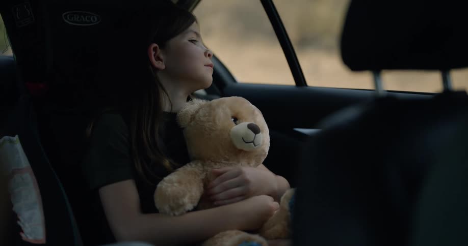 A child is riding in back of the car holding a teddy bear