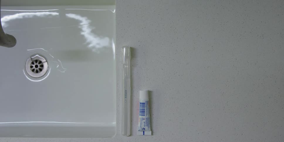 A bathroom sink with a toothbrush and toothpaste on the counter with a glass and someone fills the glass with water