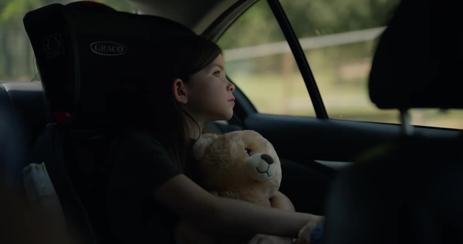 Girl with black shirt holds teddy bear and sits in safety car chair yawning