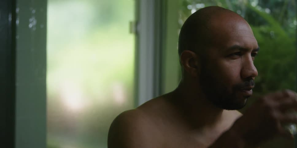 A bare chested man is standing near a window and he takes a drink from a glass of water and tips his head back