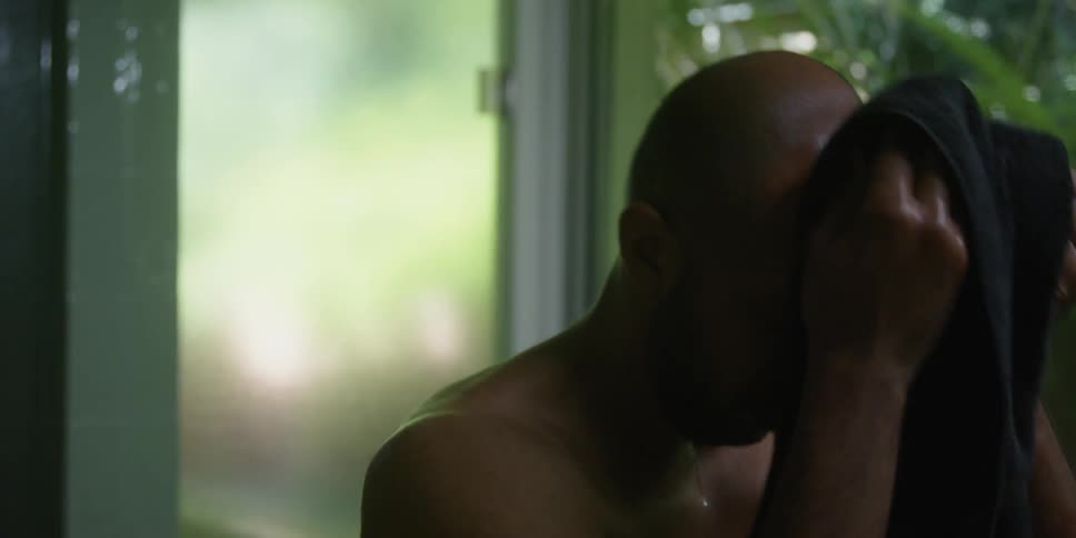 While man dries his face with a towel another persons approach and retreat is reflected in nearby glass
