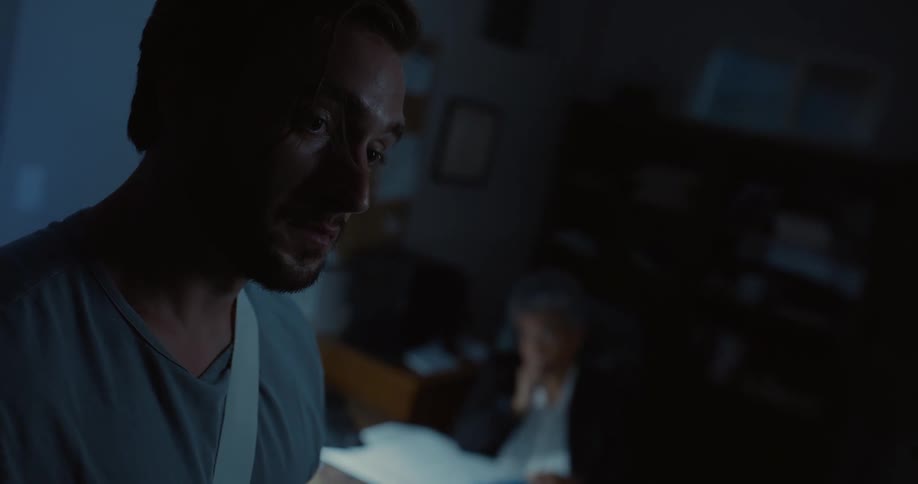 Man sitting on a desk in a dark office looks at an injured man standing in front of him