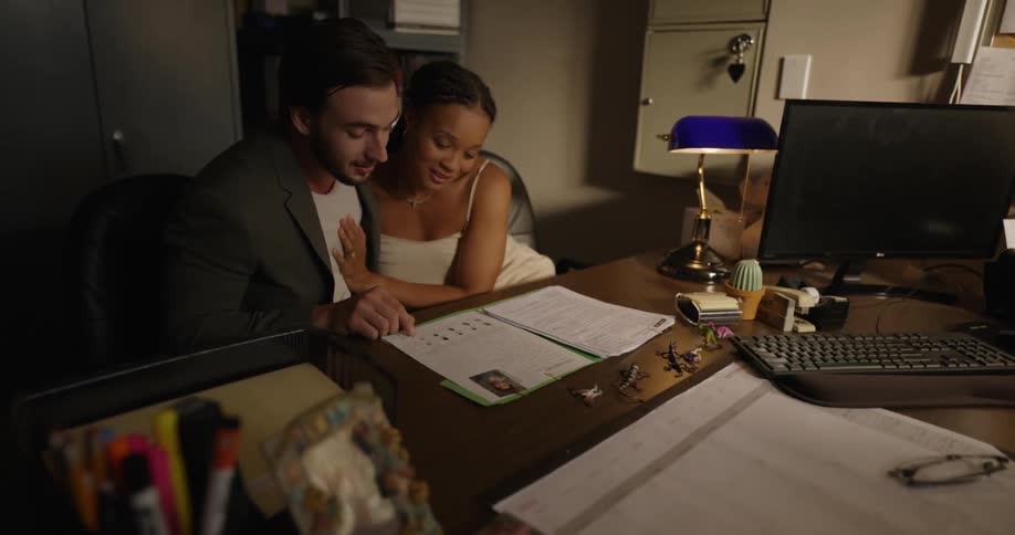A woman in an office sits across the desk from a couple looking a folder and talking while the woman rubs his chest