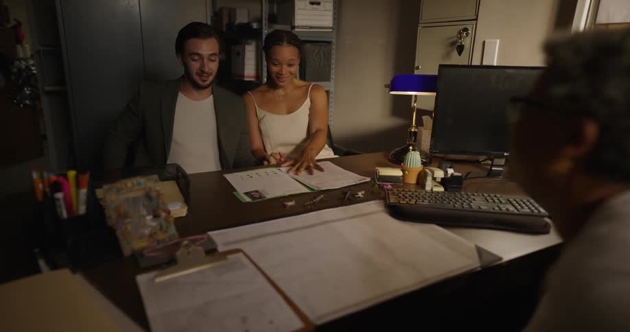 A woman carries a folder over to a nervous couple sitting at her desk sharing a photo of their new daughter