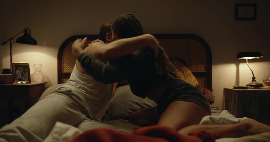 Man with beard white undershirt and gray pants makes out with woman with dreadlocks black shirt and shorts on bed in bedroom