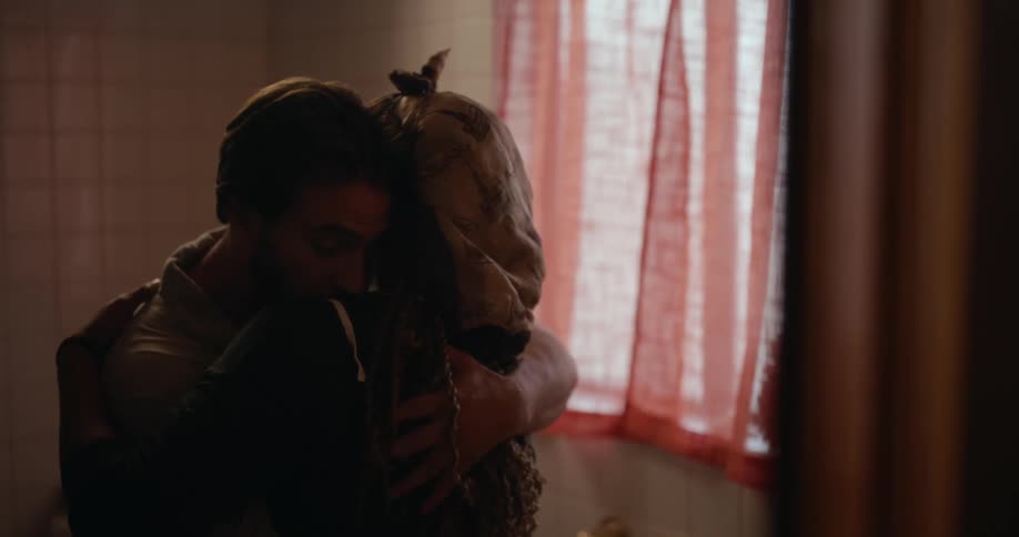 A man and a woman have their arms around each other and are kissing in a room near a window with curtains