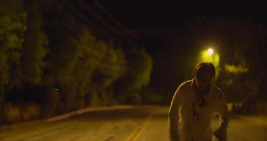 An injured man is running down a street at night with blood on his face and shirt then he slows down and collapses