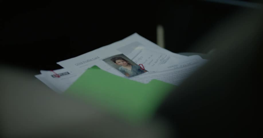 A woman sitting in a moving car reaches over and picks up a photo of a child from a folder of papers