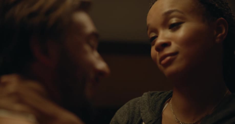 Man with beard white undershirt and gray pants makes out with woman with dreadlocks black shirt and shorts on bed in bedroom