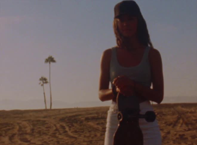 A teenage girl wearing a cap is standing on a beach with her skateboard and looking around