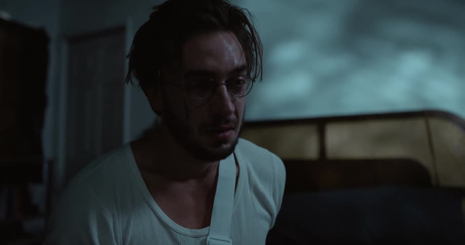 Sad man with beard white shirt jeans and broken arm sits on bed