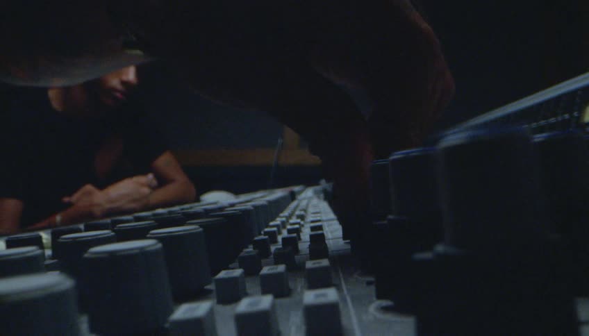 A man is turning knobs on a soundboard as another man sits near him