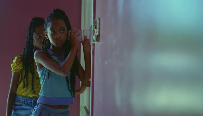 A girl holding a glass to a door listening to what is going on with her sister standing behind her