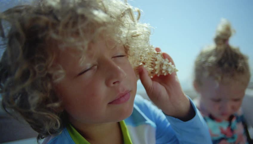 Blonde boy with curly hair holds a conch to his ear next to his sister