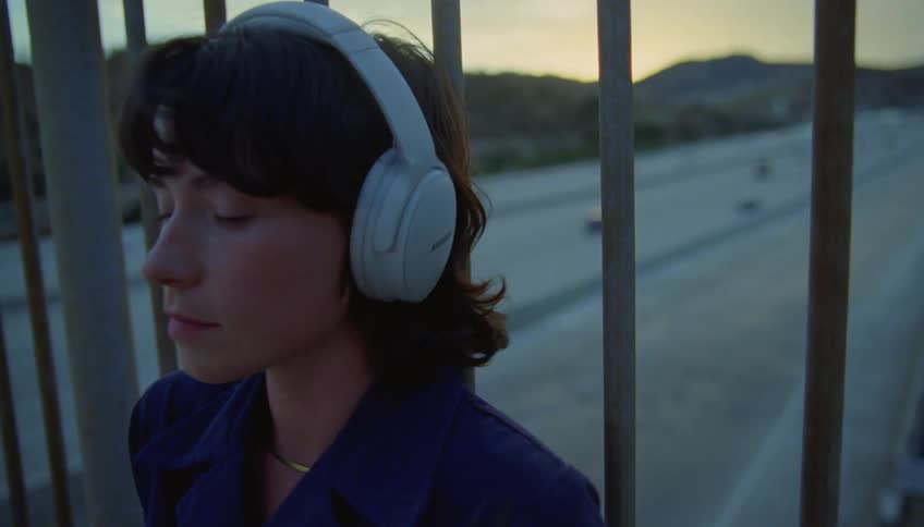 Woman leans against bars listening to music on headphones as traffic flows along controlled access highway in the background