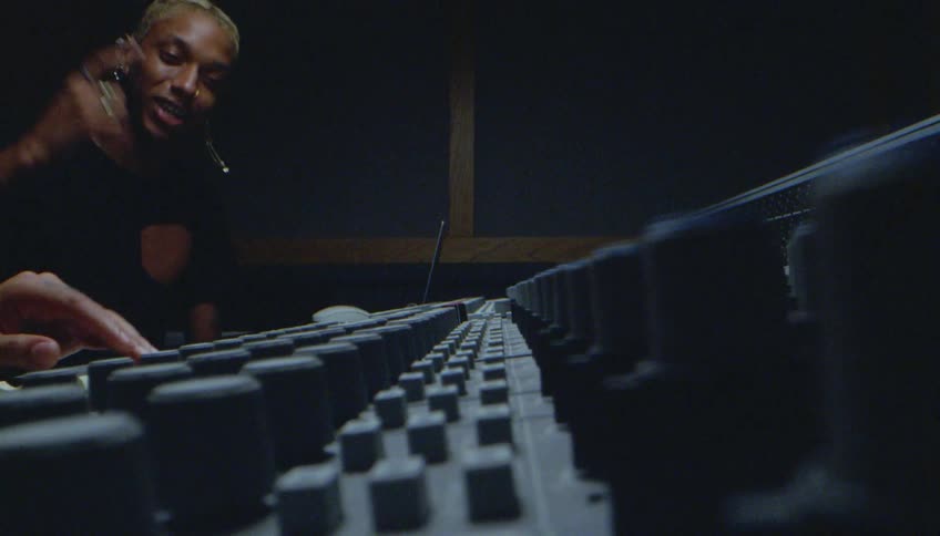 A man reaches to adjust music recording equipment as a person nods keeping the beat