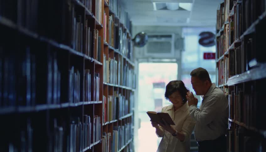 A woman is standing in a library holding an open book as a man standing next to her whispers in her ear and points at the book
