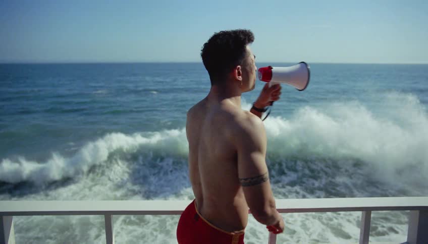 Man standing near rail watches large waves crash and makes announcement over megaphone