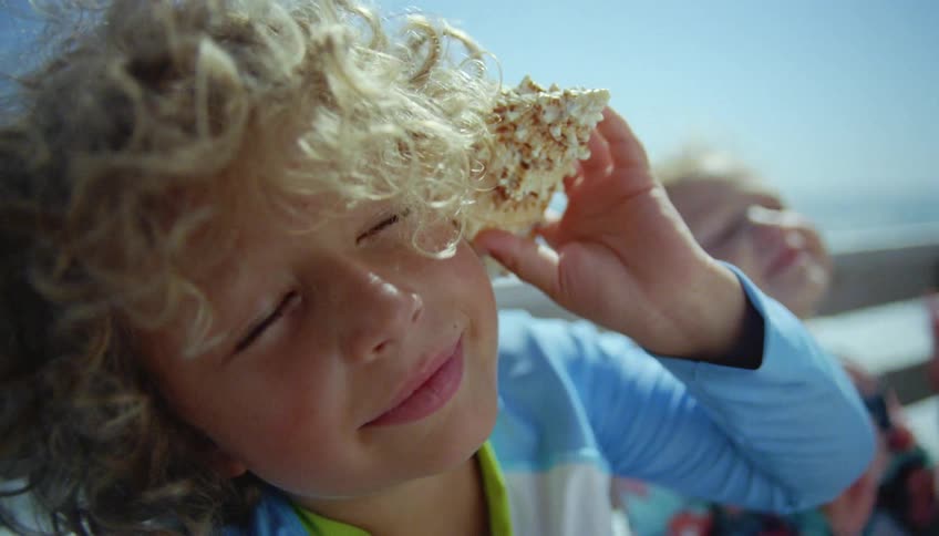 Two children are sitting on a beach and a little boy is holding a sea shell to his ear