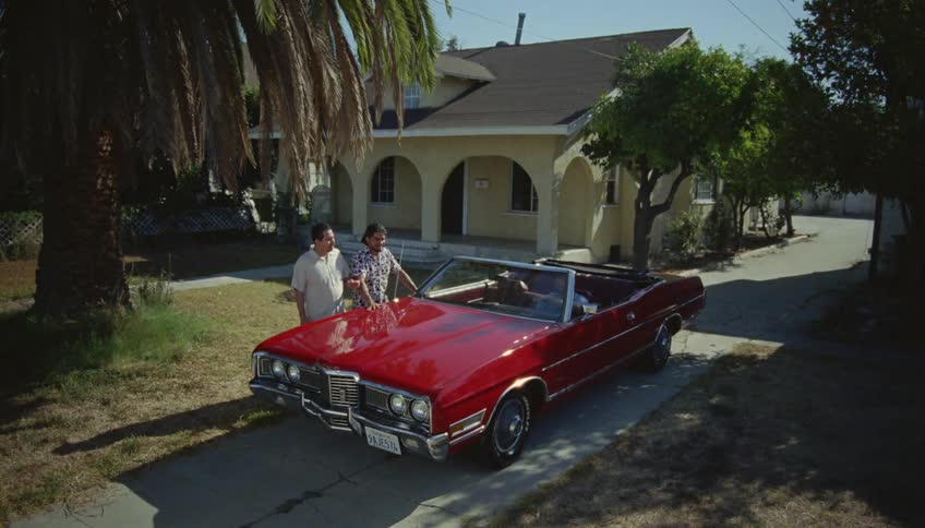 Two men stand talking with car driver sitting in driveway in a classic red convertible