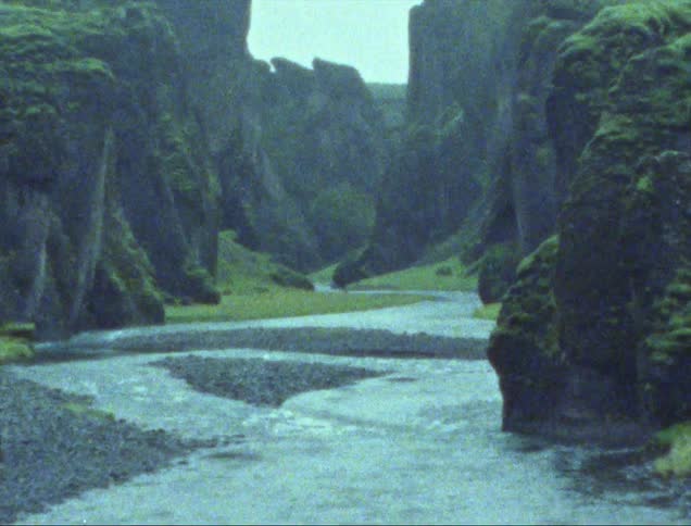 River flows around pebble strewn sandbars and between vegetation covered cliffs as people move across plateau near the edge