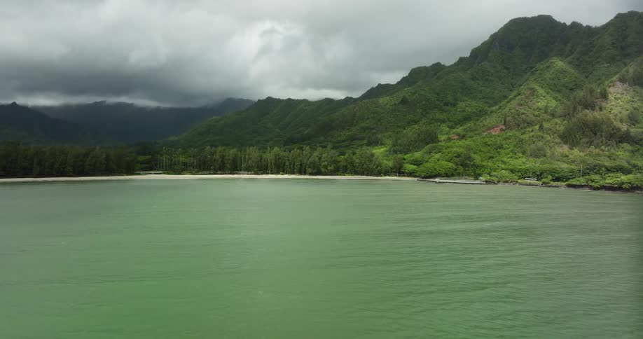 Traffic moves on a road alongside a lake and a forest with mountains in the distance under a cloudy sky