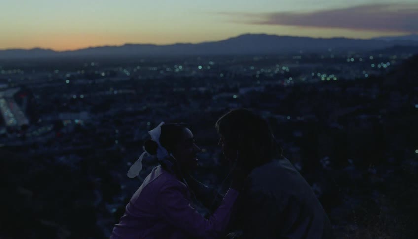 Two teenagers kiss on a hill with the city skyline below them