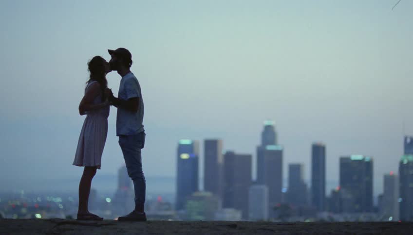 Couple partly silhouetted against the sky on a hazy day kiss with the city skyline in the background