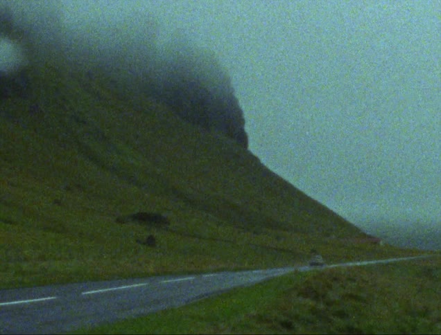 An automobile travels on a mountainside road with rock formations on top of the mountain