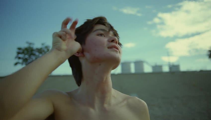 A bare chested teenage boy is standing outside and he makes a gesture with his hand