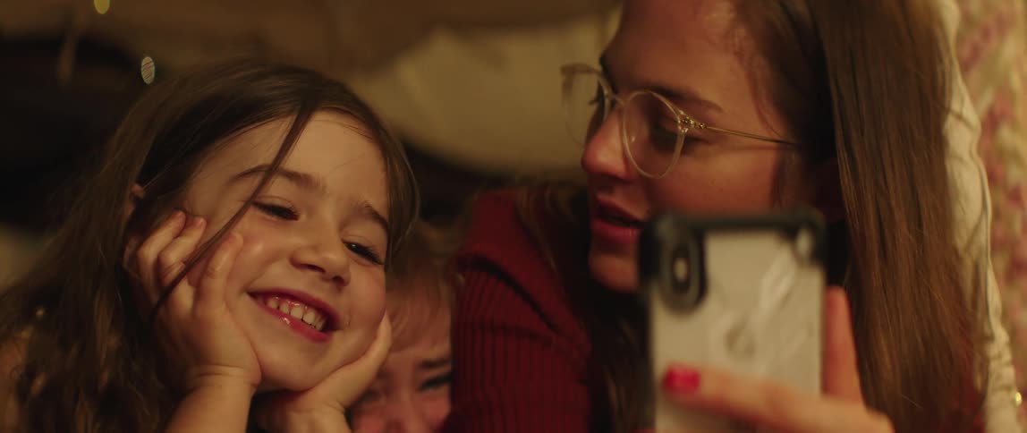 A woman holds her phone up in front of her two daughters one smiles and the other cries