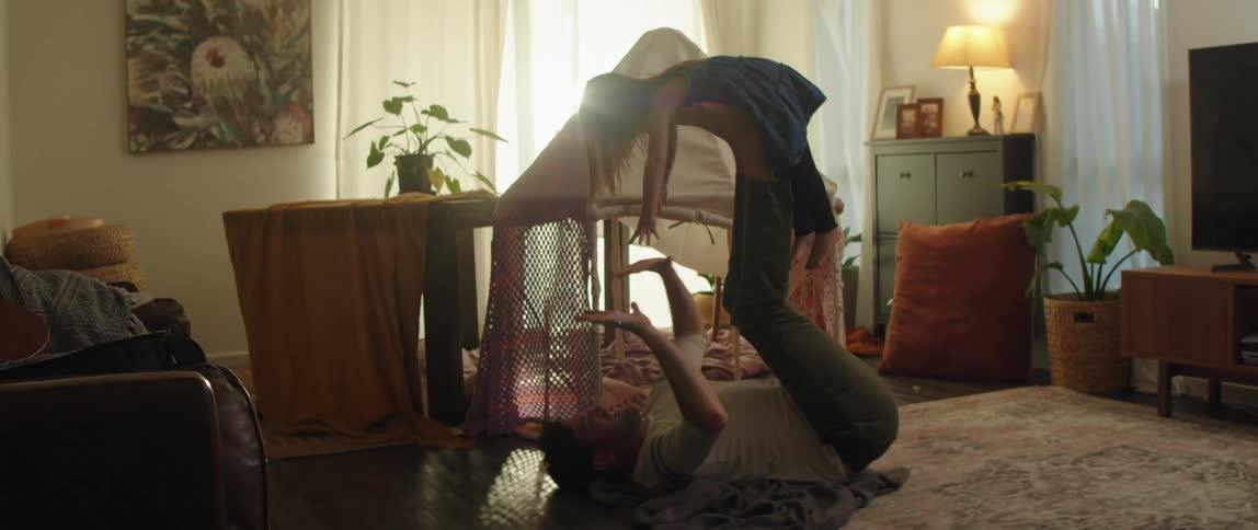 Father with white sweatshirt and jeans lifts up daughter with his feet in living room