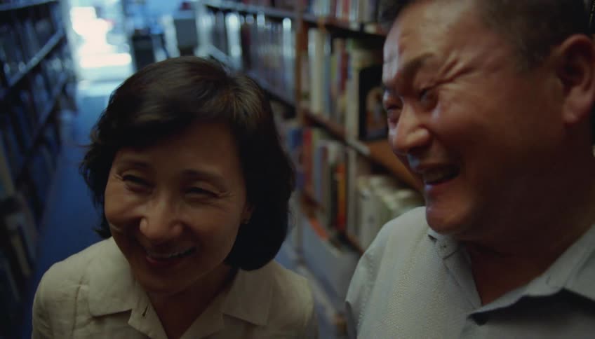 Man whispers into womans ear and they both smile as they stand between long shelves of books