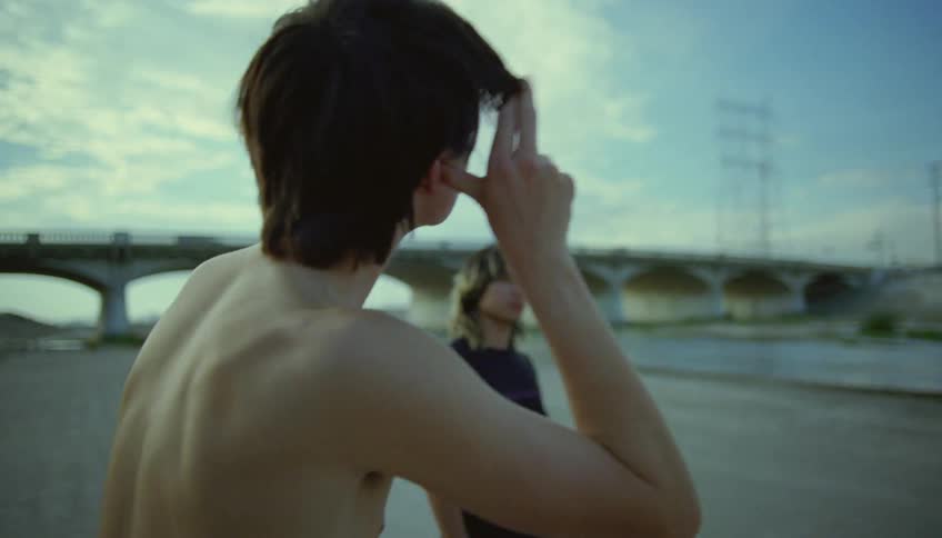 Two boys stand by a river and one boy puts his fingers by his eye and waves them at the other boy