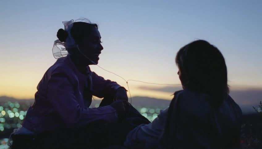 Friends sit together sharing earbuds with city behind them stretching to the mountains