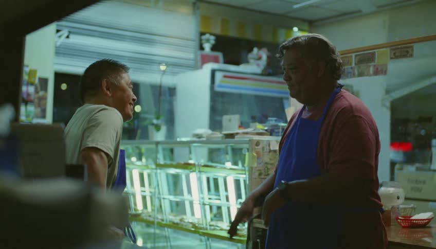 Two men wearing aprons are standing inside a restaurant talking and one man is moving his hands as he talks