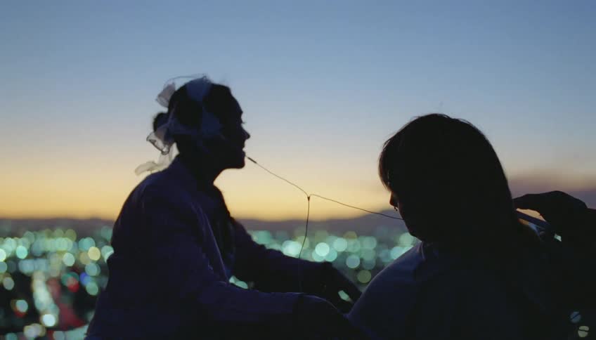 A teenage boy lies on the ground above the city skyline next to a girl who taps his chest as they both listen to music through shared headphones
