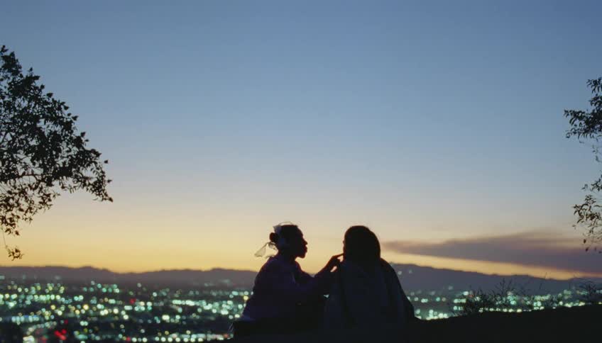 Silhouette of two people sitting and talking as the city lights shine below with mountains in the distance