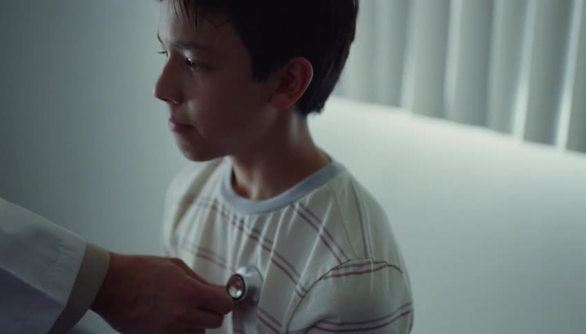 A boy is sitting in a doctors office as the doctor puts his stethoscope up to his chest