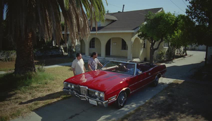 Men talk around a vintage red car parked in the driveway of a house