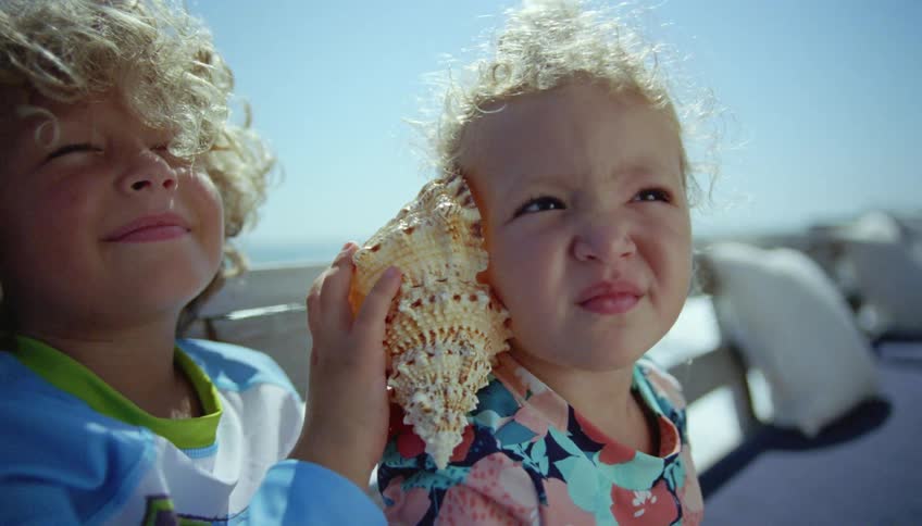 Boy holds seashell to girls ear as they sit overlooking water on sunny day