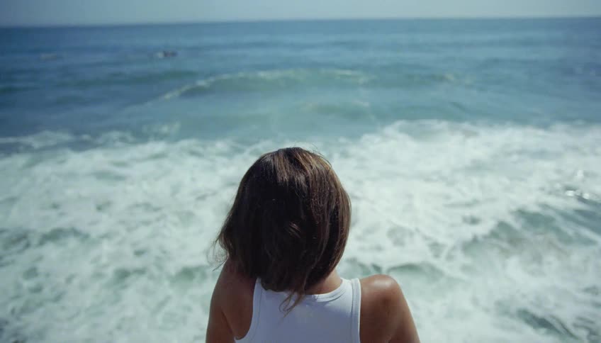 Woman with wavy hair and white top watches sea waves breaking against the coast