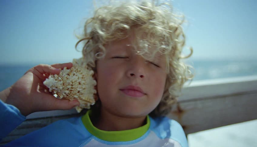 Blonde boy with curly hair holds a conch to his ear next to his sister on a wooden pier
