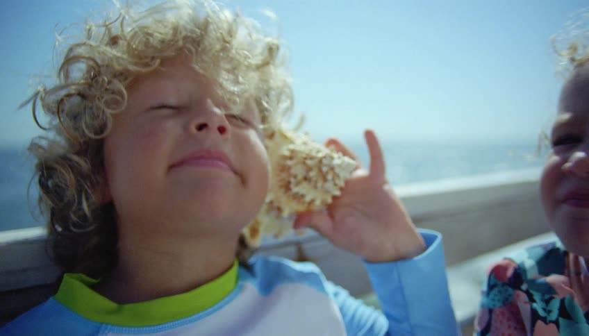Blonde boy with curly hair holds a conch to his sister ear on a wooden pier next to the ocean