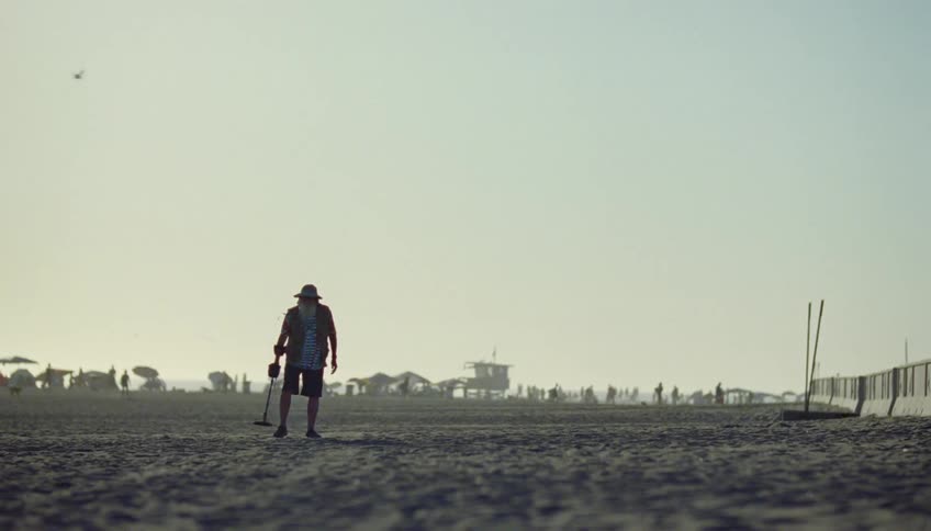 A man searches the sand on a beach with a metal detector while lots of people walk on the beach behind him